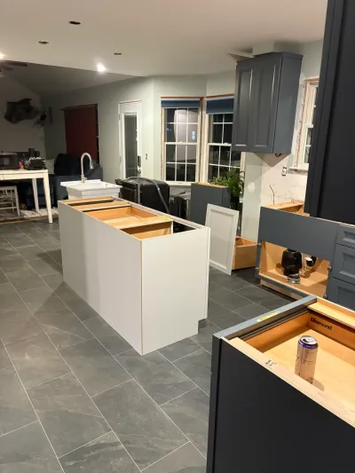Kitchen under installation with white island cabinet boxes, dark shaker cabinets, and gray tile floor.