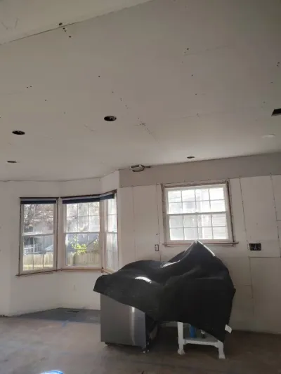 Kitchen under renovation with unpainted drywall, bay window, recessed light rough-ins and plastic-covered island.