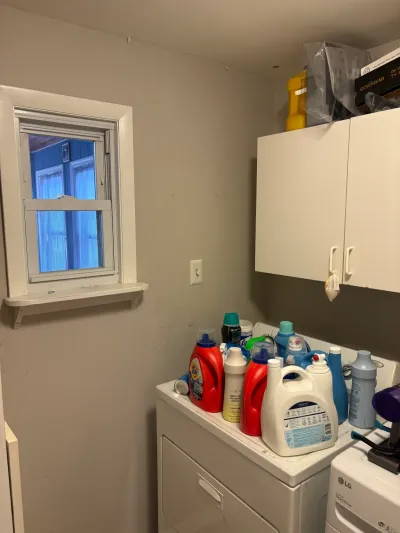 Small beige laundry room with a white wall cabinet, dryer covered in detergent bottles, and a white window.