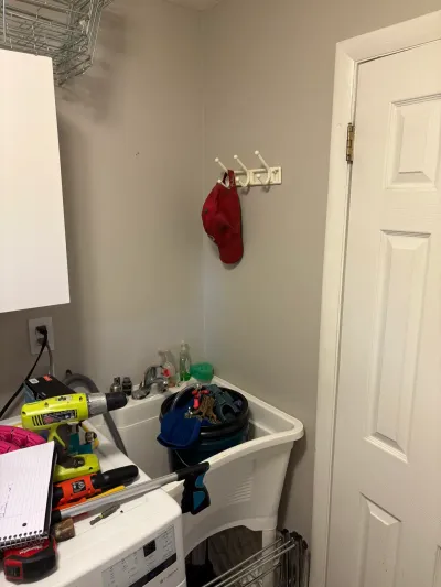 Narrow laundry room corner with white plastic utility sink, chrome faucet, and tools staged on a washer top.
