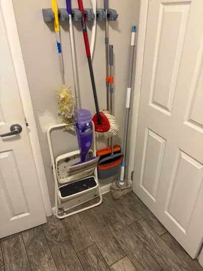 Corner nook with wall-mounted rack holding brooms and mops above a folding step stool on wood-look tile.