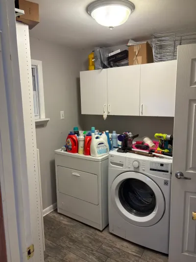Narrow laundry room with white front-load washer and dryer, white upper cabinets, and wood-look tile floor