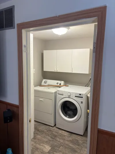 Small laundry room with white washer and dryer beneath white wall cabinets, seen through a wood-trimmed doorway.