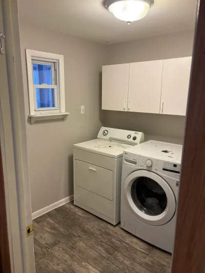 Small laundry room with white washer and dryer, white upper cabinets, and gray wood-look tile floor.