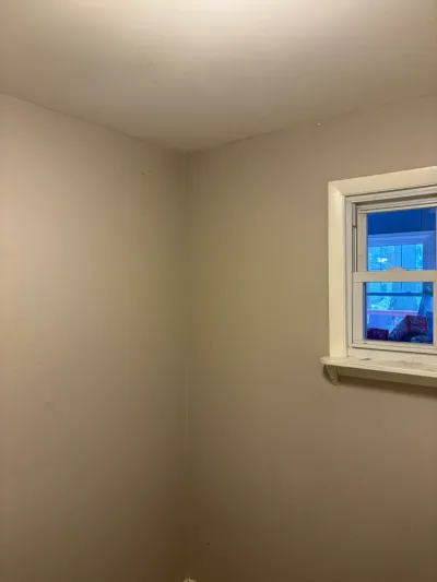 Empty beige-walled room corner with a single white-trimmed double-hung window before remodeling.