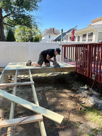 Worker kneeling with a circular saw on a new pressure-treated deck platform beside an existing red deck.