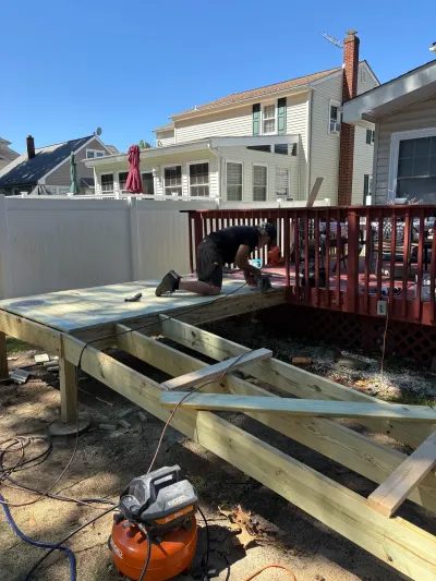 Worker kneeling on a partially built pressure-treated deck beside an existing red deck in a sunny backyard.
