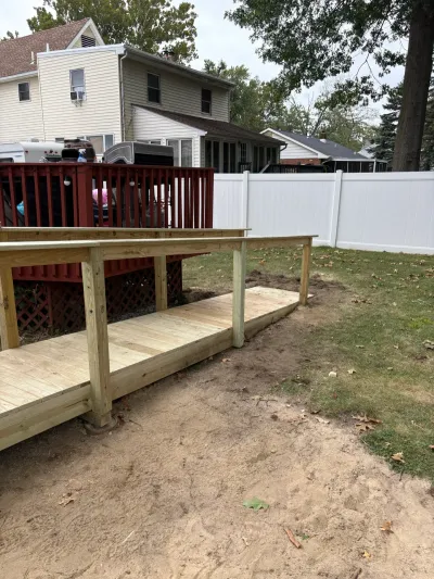 New pressure-treated wood deck and ramp built off an existing red-stained raised deck in a fenced backyard.