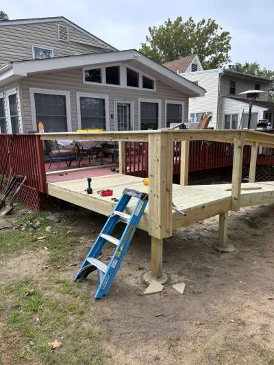 Partially built pressure-treated deck with guardrail posts and a ladder beside an older red-stained deck.