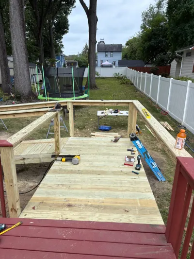 New pressure-treated deck platform under construction with railing posts, tools and a fenced backyard beyond.
