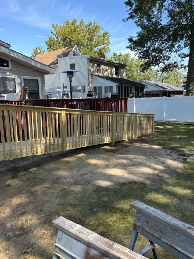 New pressure-treated deck and railing under construction beside an older dark red stained deck in a sunny backyard.