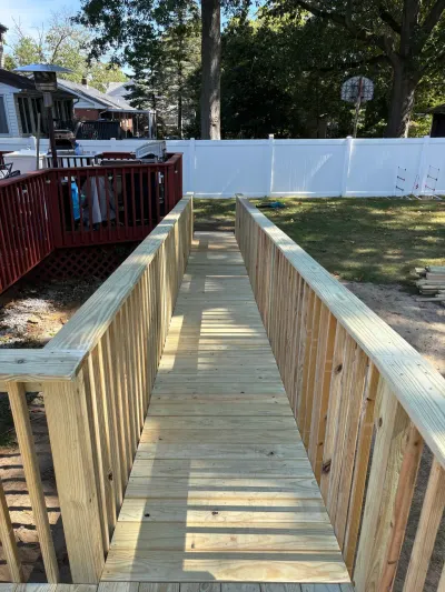 New unfinished pressure-treated wood ramp with wood guardrails running from a red stained deck into a fenced backyard.