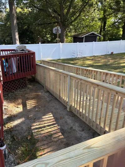 Newly framed pressure-treated deck ramp with wood railings beside an older red deck in a fenced backyard.