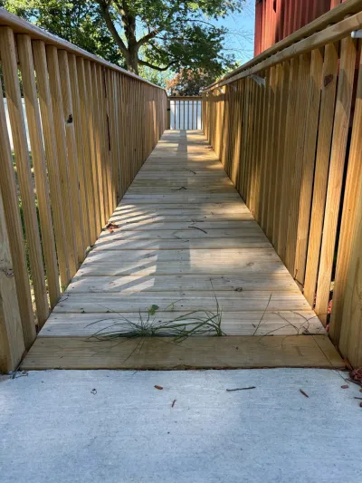 Pressure-treated wood ramp with picket guardrails rising from a concrete slab beside a red-sided house.