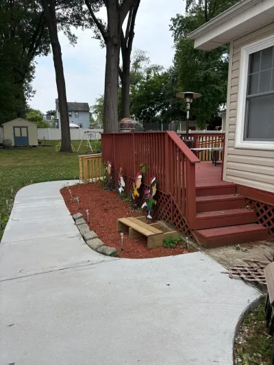Red-stained backyard deck with stairs and lattice skirting beside a new curved concrete walkway and mulch bed.