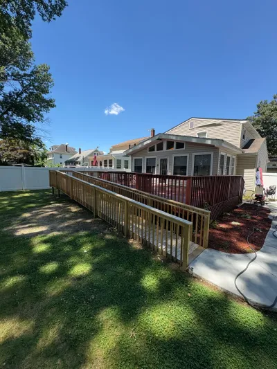 Long wood accessibility ramp leading to a red-stained rear deck on a tan vinyl-sided house.