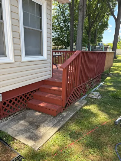 Existing red-stained wood deck with lattice skirting and stairs down to a concrete pad beside a beige-sided house.