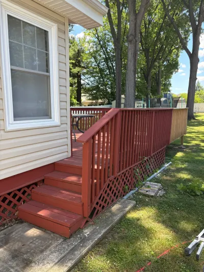 Red-painted backyard deck with lattice skirting and steps beside tan vinyl siding, unpainted railing section at right.