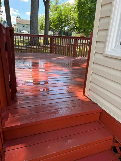 Freshly stained red wood deck with railing and steps, still wet, beside cream vinyl siding on a sunny day