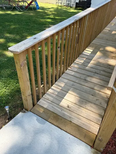 Newly built pressure-treated wood deck walkway with baluster railing meeting a concrete slab beside a lawn.