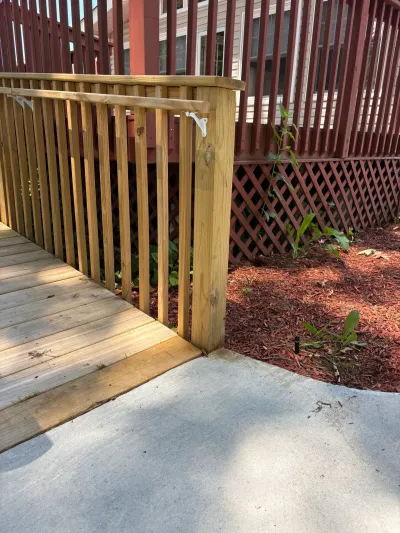 New pressure-treated wood ramp with baluster guardrail meeting a concrete patio beside a red mulch bed.