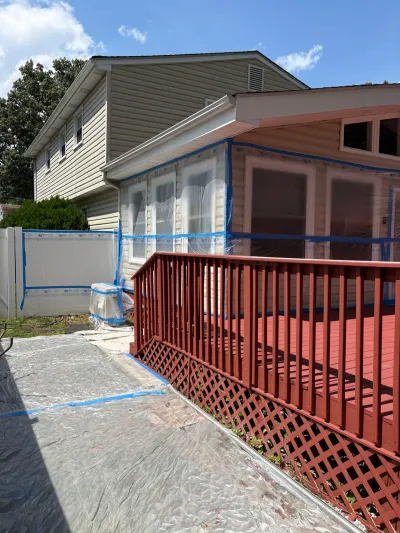 Beige-sided house with windows masked in plastic and tape beside a red painted deck during exterior prep work.