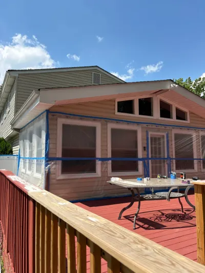 Home exterior mid-repaint in salmon pink with windows masked in plastic, viewed from a red-stained deck.