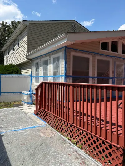 Tan-sided house with addition masked in plastic and blue tape beside a red-stained deck during exterior prep.