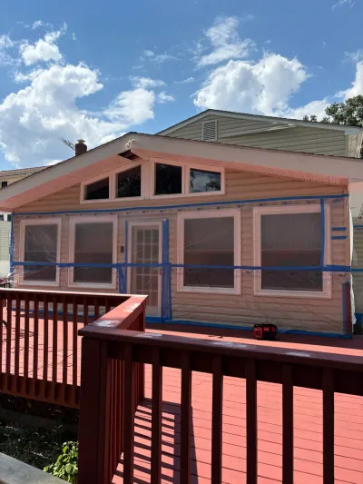 Pink-painted house exterior with windows masked in plastic and blue tape, seen from a red wood deck.