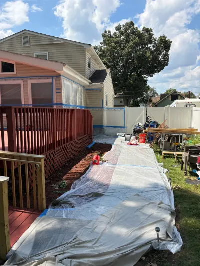 Home exterior masked with plastic and tape during repainting, tarps covering lawn beside a red-stained deck