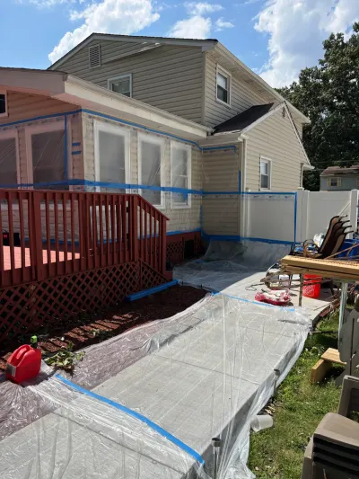 Two-story tan-sided house with siding, windows and patio masked in plastic sheeting and blue tape.