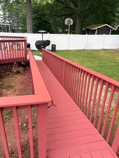 Red-painted backyard deck with an attached access ramp, wood railings and handrail, beside a white vinyl fence.