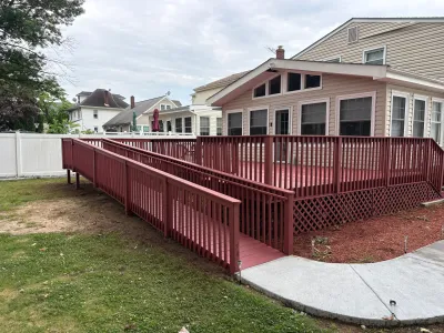 Barn-red wood deck with a long switchback wheelchair ramp leading to a concrete walkway in a backyard.