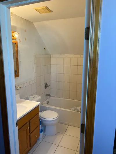 Dated small bathroom with white tile tub surround, oak vanity, and sloped ceiling before remodeling