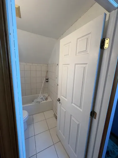 Small dated bathroom with white square tile, alcove tub and toilet under a sloped ceiling, door open.