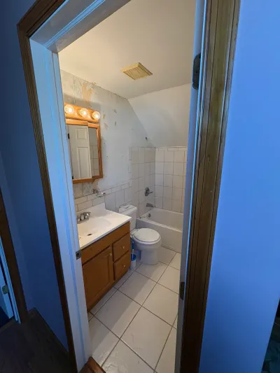 Dated small bathroom with oak vanity, white tile tub surround, torn wallpaper, and sloped ceiling over the tub.
