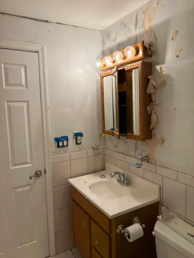 Dated bathroom with oak vanity, tri-view mirror cabinet, white tile wainscot and peeling wallpaper.