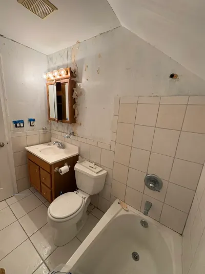 Dated bathroom before remodeling with sloped ceiling, white tile, oak vanity, and peeling wallpaper.