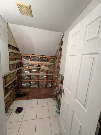 Bathroom stripped to studs during demolition, with white tile floor, sloped ceiling and open panel door.