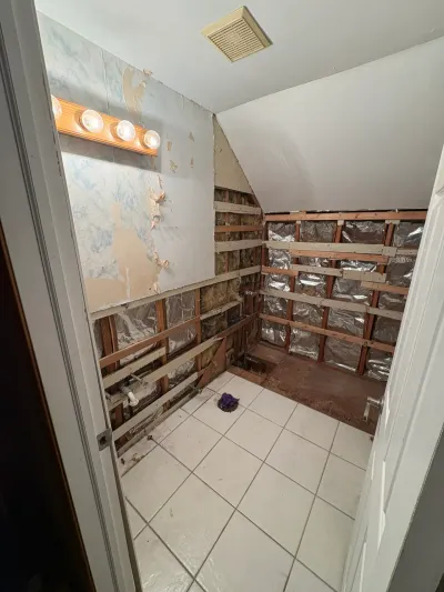 Small bathroom stripped to studs during demolition, with tile floor intact and a sloped kneewall ceiling.