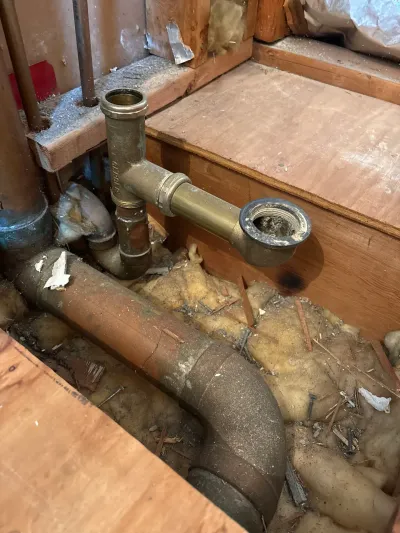 Exposed brass tub drain trap and cast iron waste pipe in an opened bathroom floor cavity during demolition.