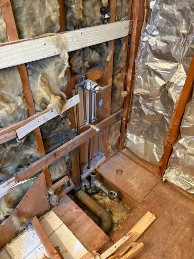 Bathroom stripped to studs with exposed insulation, chrome shower valve and open floor showing drain piping.