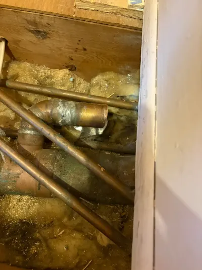 Copper and cast iron pipes exposed in an opened floor cavity filled with fiberglass insulation and sawdust.