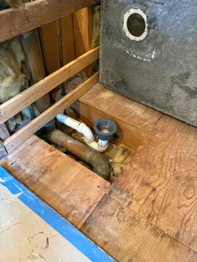 Shower drain rough-in with PVC trap and cast iron line exposed through cut-open plywood subfloor