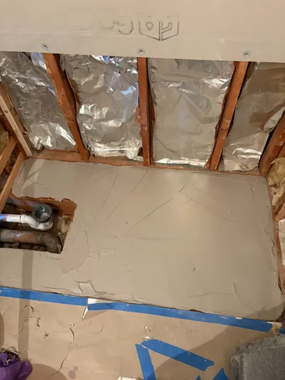 Bathroom subfloor coated in gray leveling compound with a plumbing access cutout and insulated open stud wall behind.