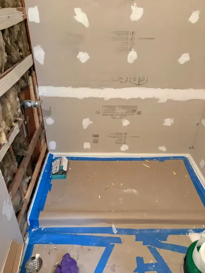 Bathroom alcove mid-remodel with fresh drywall, open insulated stud wall and taped floor protection.