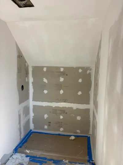 Small bathroom under construction with taped drywall, gray backer board alcove, sloped ceiling and papered floor.