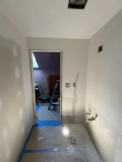 Bathroom under renovation with bare drywall, capped plumbing stubs, taped floor protection and a work light.