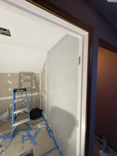 In-progress room with sloped ceiling, patched drywall, taped subfloor and a step ladder seen through a doorway.