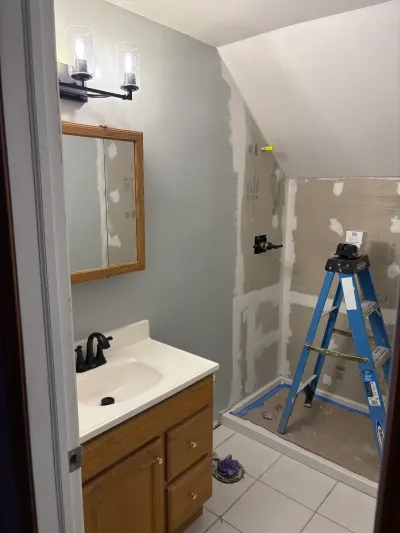 Bathroom under renovation with oak vanity, gray walls, and a shower alcove stripped to backer board under a sloped ceiling.